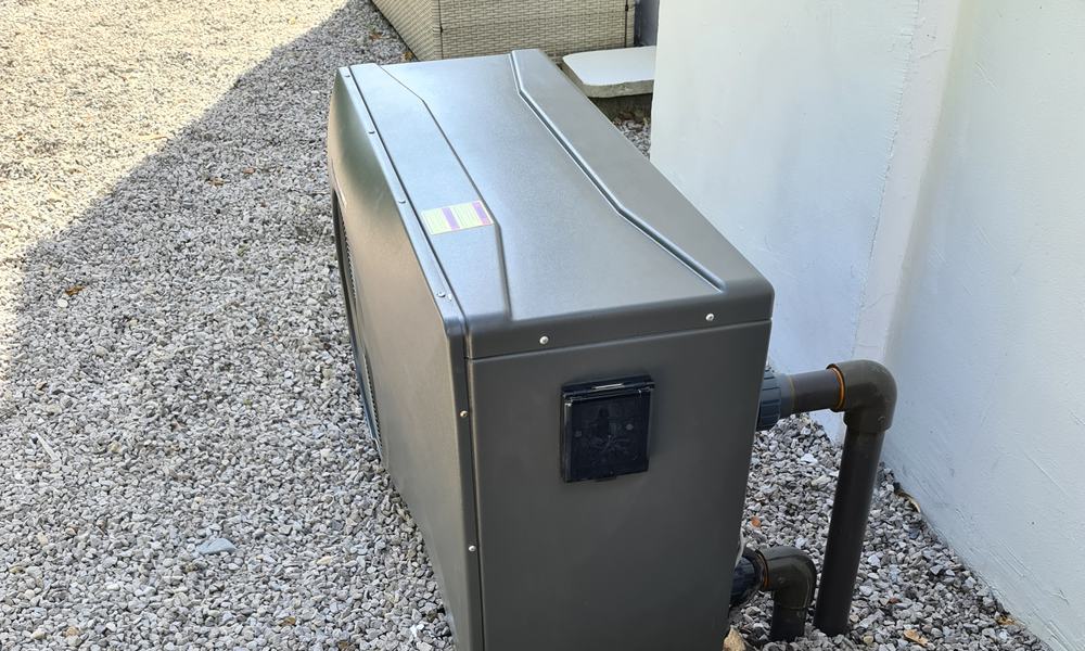 Pompe à chaleur de piscine posée sur gravier contre un mur, avec ses raccords hydrauliques
